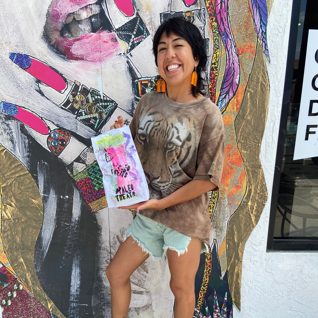 Small stack sampler smiling woman holds up sampler bag of paleo treats outside san diego store.