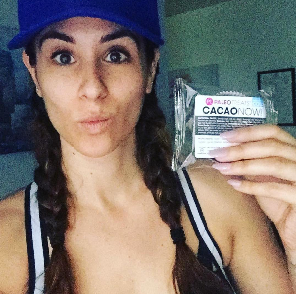 Smiling dark haired woman with braids holding up cacao now paleo treats chocolate bar.