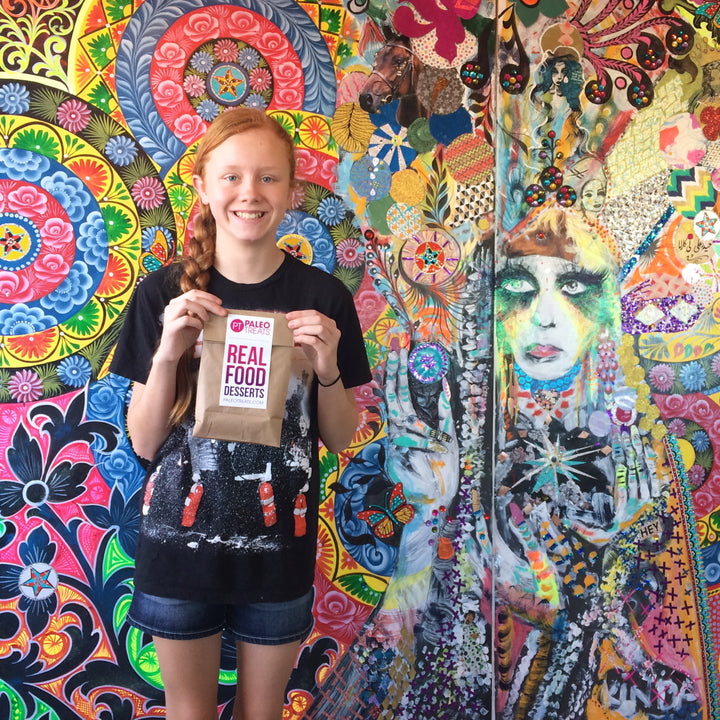 Smiling female child holding up bag of paleo treats with real food desserts sticker.